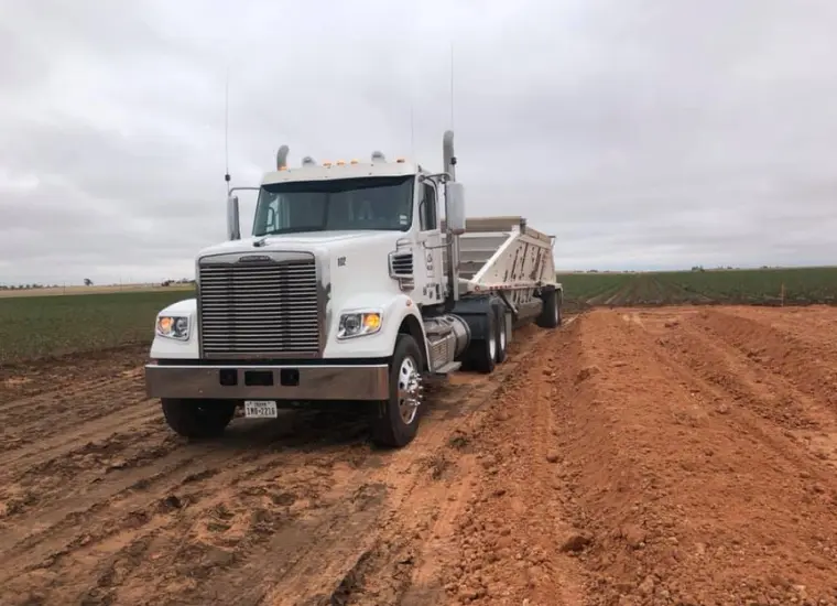 A row of five heavy construction and farming vehicles, including rollers, a grader, and a tractor, are lined up on a vast, flat dirt field under a cloudy sky during daylight. A large white semi-truck with a trailer is parked on a dirt road next to plowed farmland under a cloudy sky. The truck's headlights are on and the surrounding landscape is mostly flat with fields extending into the distance.